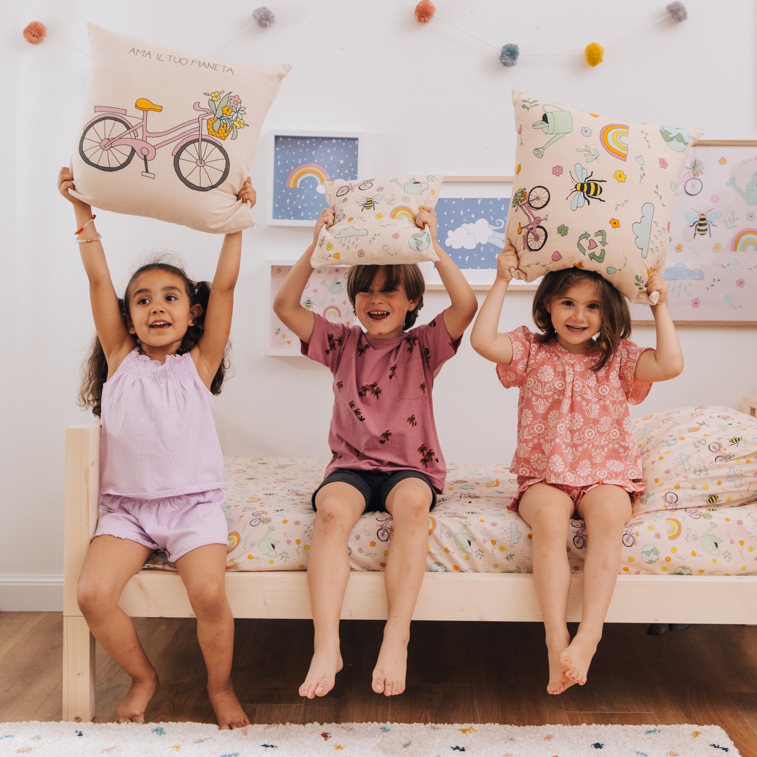 3 bambini con in mano un cuscino Ama il tuo pianeta ciascuno, che alzano sopra la testa, seduti su un letto zero+Earth by nabè.