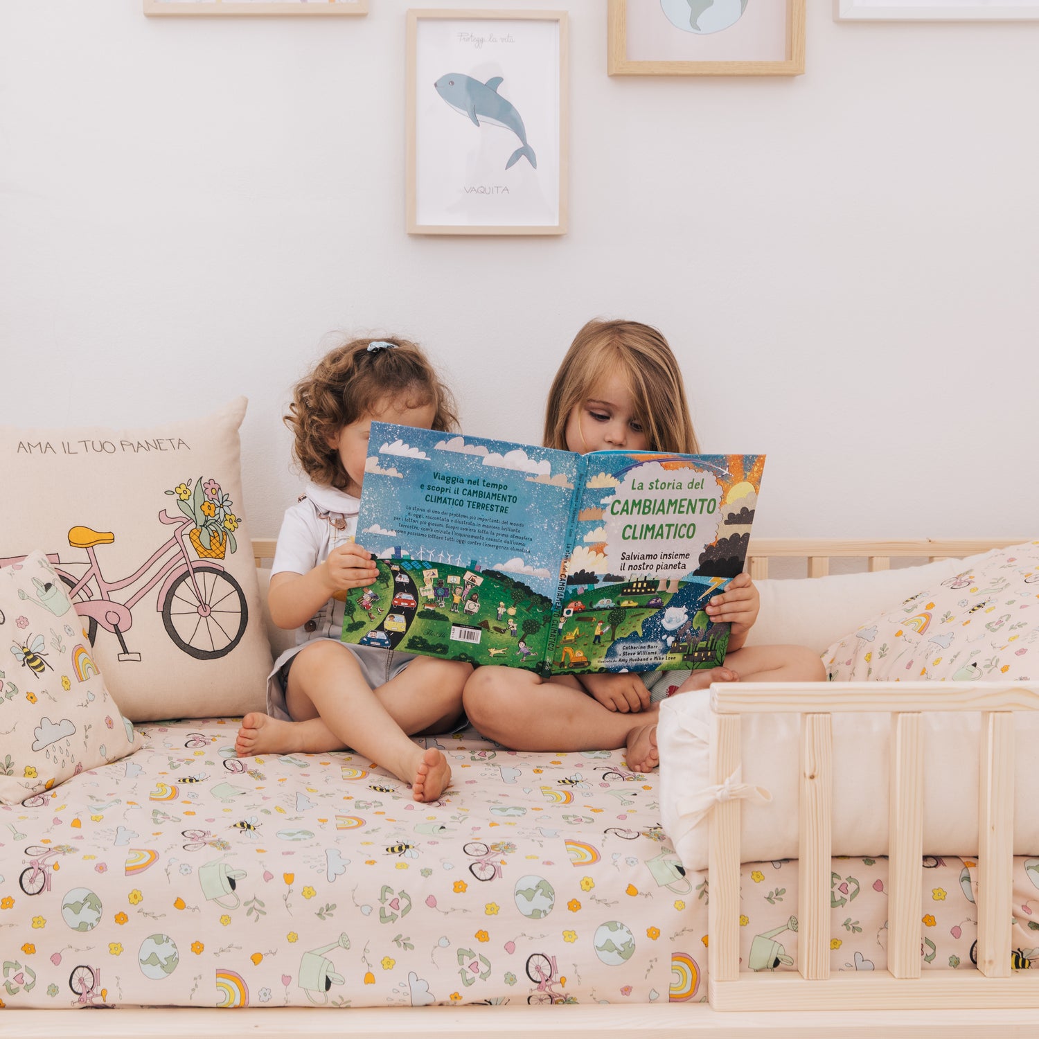 Due bambine leggono un libro sul cambiamento climatico adatto alla loro età, su un letto nabè fra cuscini a fantasia Ama il tuo pianeta.