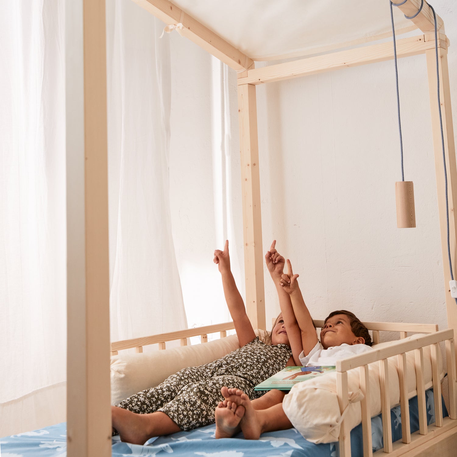 due bambini sdraiati in un lettino montessoriano in legno naturale indicano il telo per copertura baldacchino del letto nabè